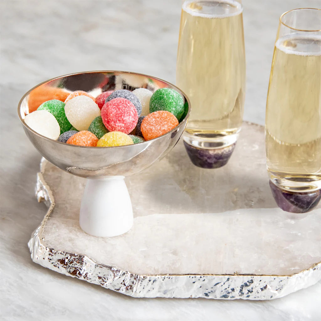 An Anna New York Coluna Carrara Marble Nut Bowl filled with colorful gumdrops sits beside two champagne flutes on a white agate serving tray.