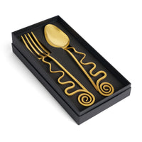 The Spirale Gold Serving Set by L'Objet comes in a luxury black gift box with a 24k gold plated spoon and fork, each featuring wavy handles ending in elegant spirals, displayed on a black foam background.