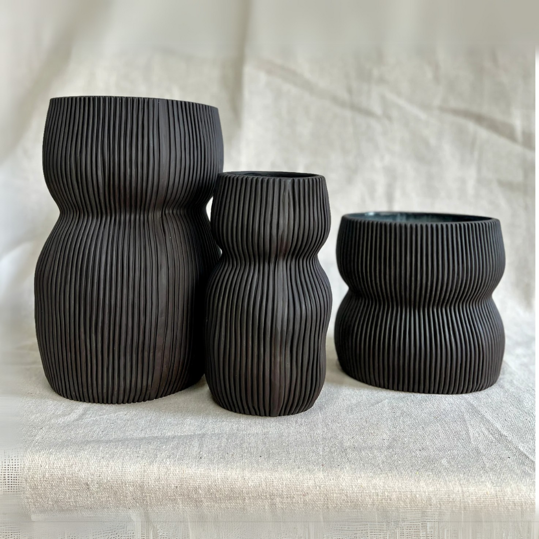 Three black Curvy Oval Vases by CYM Warkov Ceramics, each with ribbed detailing and unique heights and shapes, are arranged side by side on a light textured surface against a neutral fabric backdrop.