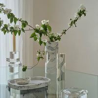 A modern glass table displays the Greg Natale Pietra Marble Vase with white flowers, a clear candle holder with a yellow candle, a marble tray and small dish, plus a silver vase with geometric accents in a bright, minimal room.