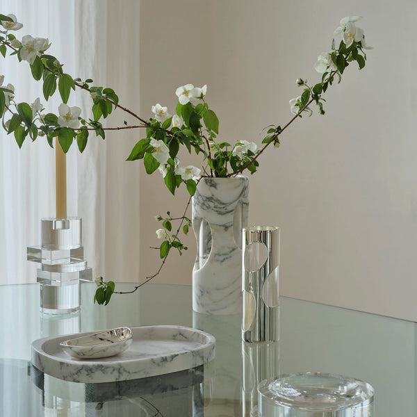 A modern glass table displays the Greg Natale Pietra Marble Vase with white flowers, a clear candle holder with a yellow candle, a marble tray and small dish, plus a silver vase with geometric accents in a bright, minimal room.