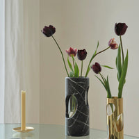 A Greg Natale Pietra Marble Vase with circular cut-out biomorphic design holds dark burgundy tulips. Beside it, a gold holder with an unlit beige candle sits on a glass table set against a light background.
