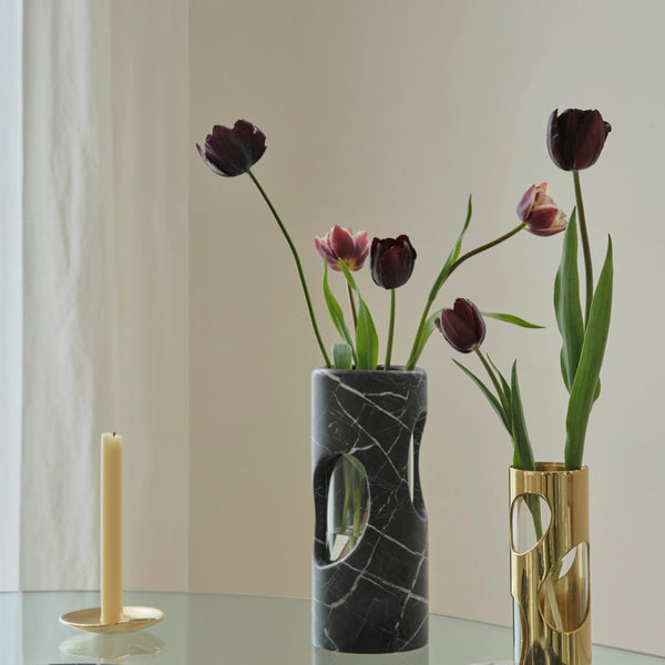 A Greg Natale Pietra Marble Vase with circular cut-out biomorphic design holds dark burgundy tulips. Beside it, a gold holder with an unlit beige candle sits on a glass table set against a light background.