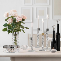 A tabletop display features the Orrefors Kosta Boda Carat Candlestick Brass Set of 2 with lit white candles, a vase of pale pink roses, modern cut glass decor, two black bottles, and a bowl against a white wall with framed art.
