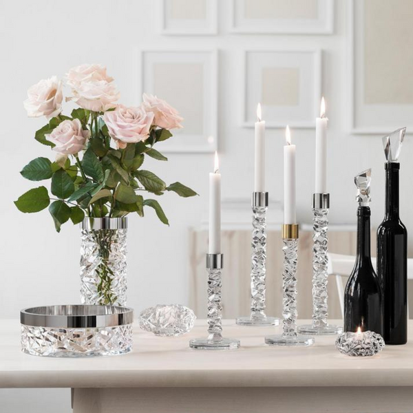A tabletop display features the Orrefors Kosta Boda Carat Candlestick Brass Set of 2 with lit white candles, a vase of pale pink roses, modern cut glass decor, two black bottles, and a bowl against a white wall with framed art.