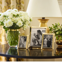 A round black table holds a vase of white flowers, a gold lamp, a green plant, and three Aerin Cecile Frames featuring an elegant three-dimensional design that displays cherished family photos. Yellow patterned curtains hang in the background.