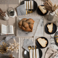 A top-down view of a table set with black plates, gold flatware, striped napkins, braided bread slices, a large loaf on the Apeloig Solid Acrylic Challah Board & Knife, glasses of water, open books, and dried florals on a neutral tablecloth.