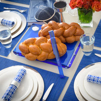 A Shabbat table set with challah bread on the Apeloig Solid Acrylic Challah Board & Knife, a kiddush cup, kippah, siddur, plates with napkins, water glasses, and orange flowers in a vase on a blue placemat.