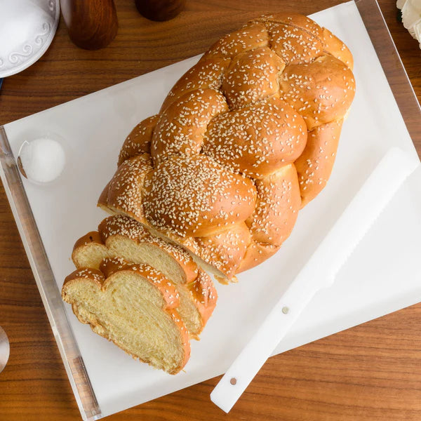 A partially sliced braided challah with sesame seeds rests on the Apeloig Solid Acrylic Challah Board, accompanied by a white serrated knife, atop a wooden table.