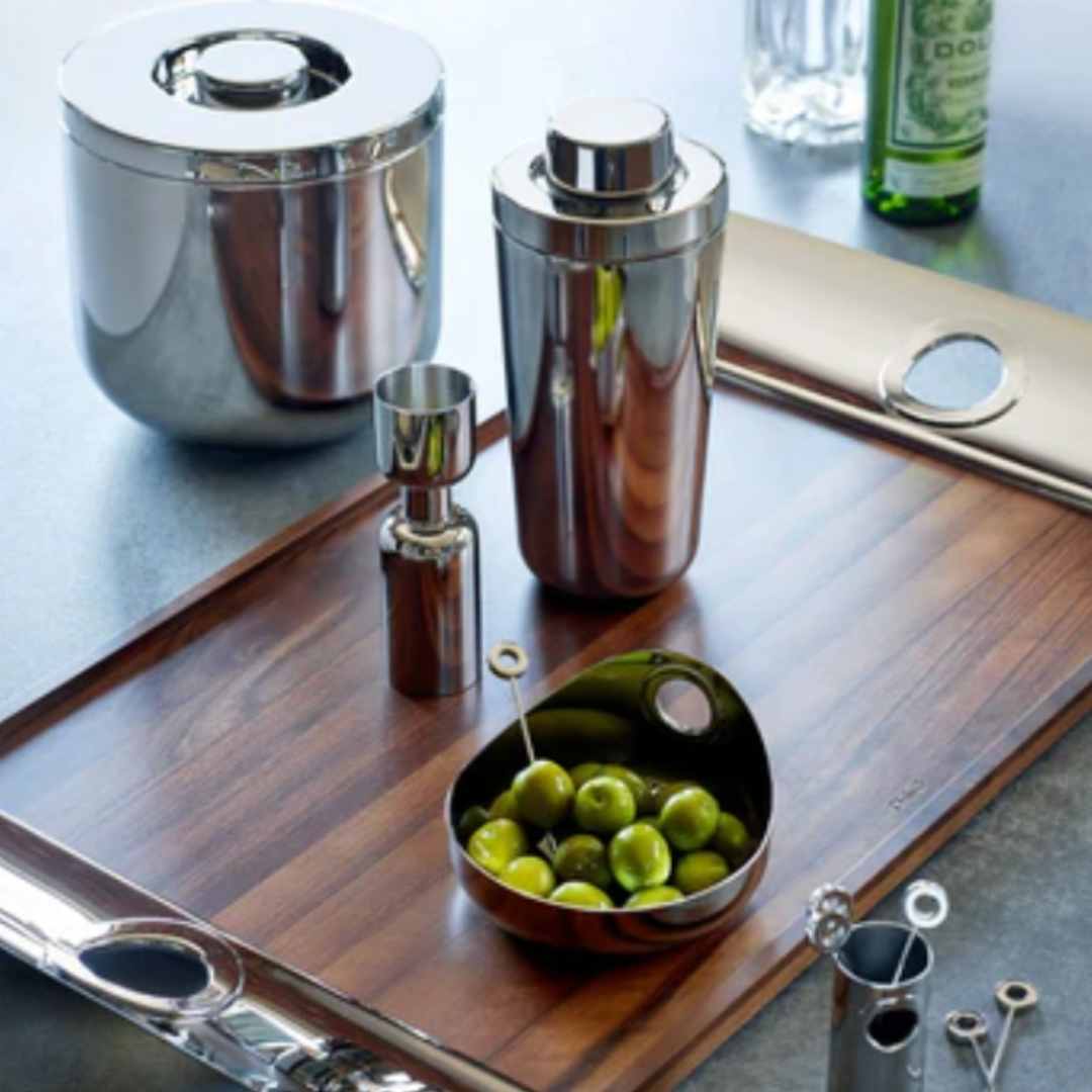 A modern wooden tray holds a Christofle Oh De Small Stainless Steel Bowl filled with green olives and picks, alongside a silver cocktail shaker, jigger, and barware. An ice bucket and bottle sit nearby on a gray surface.
