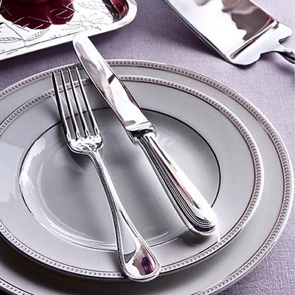 A Christofle Perles 5 Piece Setting in stainless steel rests on stacked gray plates with decorative edges atop a lavender tablecloth. A pie server and a serving tray with red food are visible in the background.