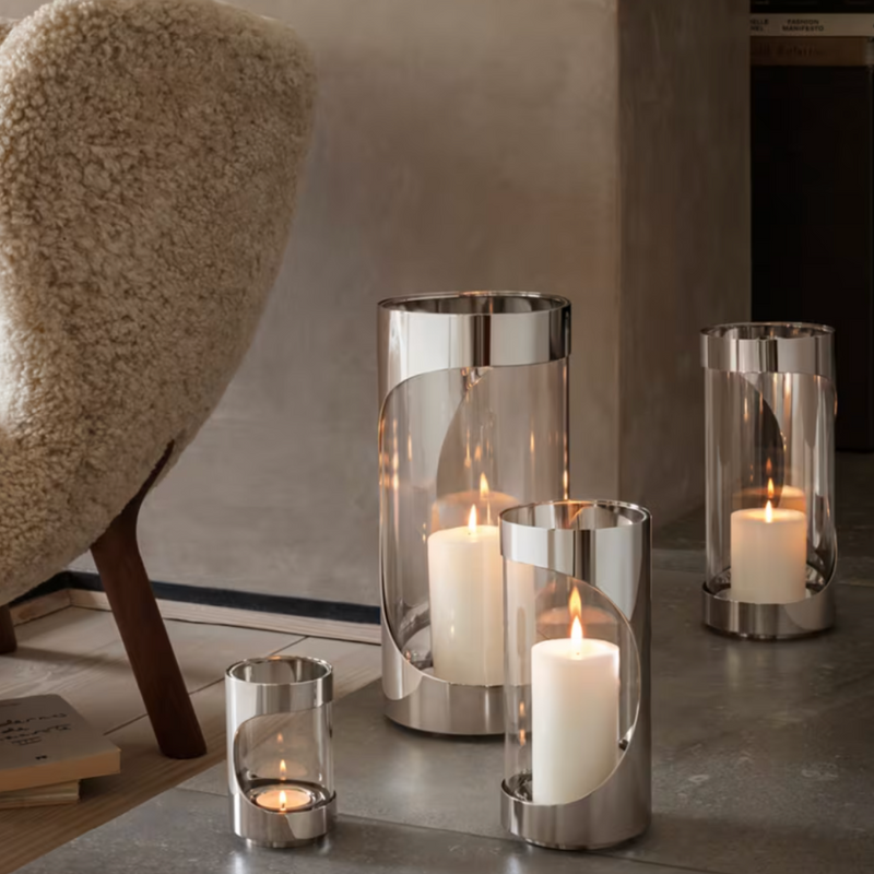 Four lit candles in glass and metal hurricane lanterns, including the Georg Jensen Chrome Contours Hurricane 13", are arranged on a stone floor beside a textured cream armchair and beige wall in this cozy modern home lighting scene.