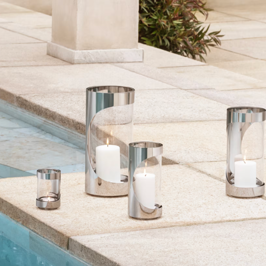 Four Georg Jensen Chrome Contours Hurricane 13" lanterns with lit white candles are arranged on a stone poolside, creating a serene and elegant outdoor setting—ideal for modern home lighting.
