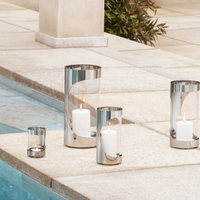 Four Georg Jensen Chrome Contours Hurricane 13" lanterns with lit white candles are arranged on a stone poolside, creating a serene and elegant outdoor setting—ideal for modern home lighting.