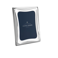 The Cobra Stainless Picture Frame by Georg Jensen is a shiny silver rectangular frame with wavy edges, standing upright and featuring a dark blue placeholder with the brand’s logo and text.