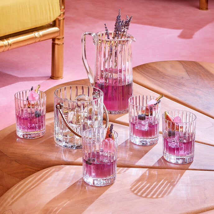 The Baccarat Harmonie Barware Set of 2 by Baccarat is displayed on a geometric wooden table, with colorful drinks garnished with flowers and fruit, set against a yellow chair and pink floor background.