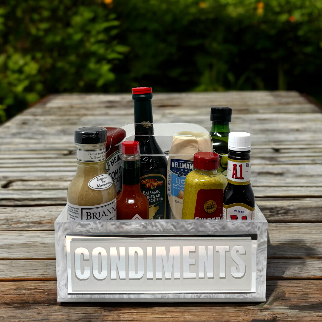 The Eye Candy Porter Acrylic Condiments Caddy sits on a wooden table, organizing sauces and dressings like mustard, mayonnaise, hot sauce, and balsamic vinegar, with greenery in the background.