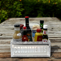 The Eye Candy Porter Acrylic Condiments Caddy sits on a wooden table, organizing sauces and dressings like mustard, mayonnaise, hot sauce, and balsamic vinegar, with greenery in the background.