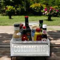 The Eye Candy Porter Acrylic Condiments Caddy keeps sauces and dressings organized on a wooden picnic table outdoors, with a garden and blooming flowers in the background.