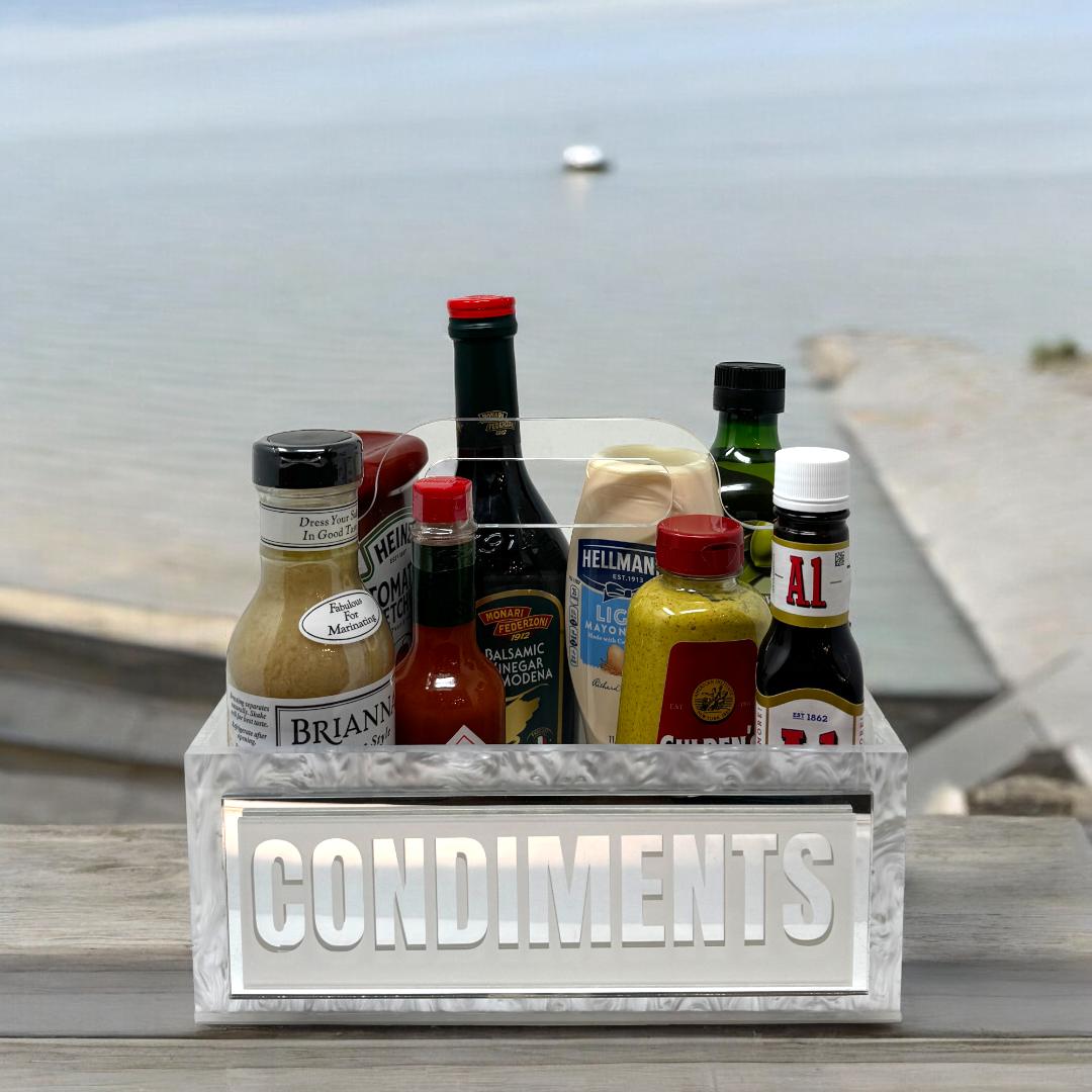 The Eye Candy Porter Acrylic Condiments Caddy, labeled "CONDIMENTS," organizes sauces and dressings on an outdoor table with a waterside view—an ideal kitchen organizer for gatherings and entertaining.