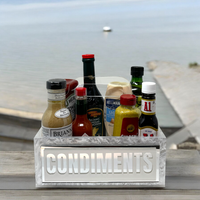 The Eye Candy Porter Acrylic Condiments Caddy, labeled "CONDIMENTS," organizes sauces and dressings on an outdoor table with a waterside view—an ideal kitchen organizer for gatherings and entertaining.