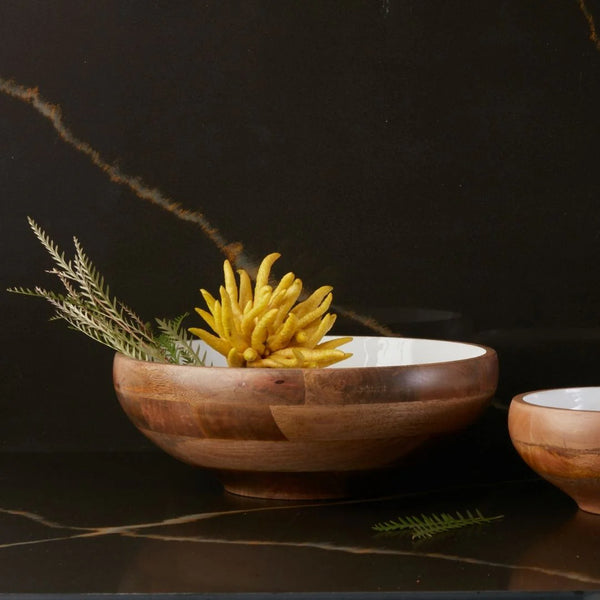 The Be Home Mayan Wood Footed Bowl - 17D" is crafted from sustainably sourced wood. It features a white interior and holds a yellow Buddha’s hand citron with green fern leaves, set against dark marble. A smaller bowl sits partially visible to the right.