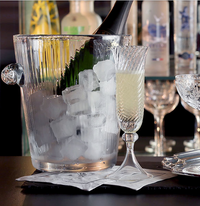 The William Yeoward Corinne Champagne Bucket chills a bottle of bubbly beside a filled champagne flute, with barware and bottles blurred in the background, evoking the glamour of the Jazz Age.