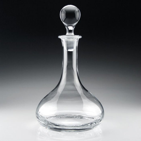 The William Yeoward Classic Ships Decanter, crafted from handmade crystal glass with a round stopper, features a wide base and narrow neck, elegantly displayed on a reflective surface against a dark gradient background.