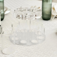 The BT-Shalom Lucite Fountain Kiddush Cup features a rotating top and multiple cylindrical slots. Its modern, clear design stands out on a white textured tablecloth, complementing green glass bottles and plates on the Shabbat table.