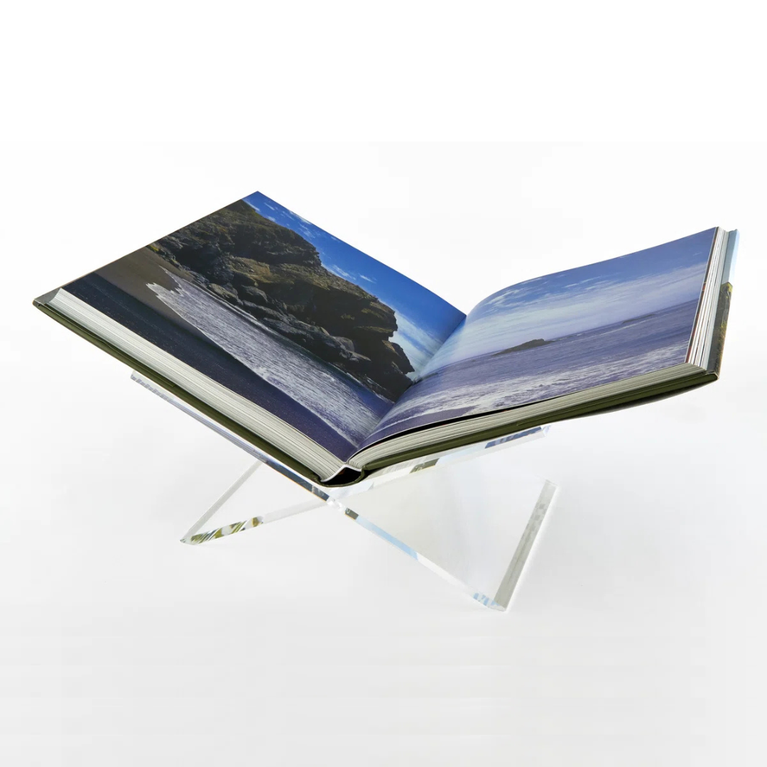 A beach photo book rests open on the Tizo Lucite Book Stand, its sleek X-shaped design offering elegant book decor against a plain white background.
