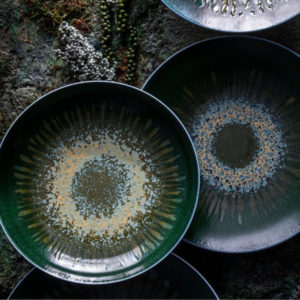 Three large Jaune De Chrome Cyclone Dinnerware plates display circular abstract patterns in blue, green, and beige, arranged on a textured mossy surface with clusters of small white and green berries nearby.