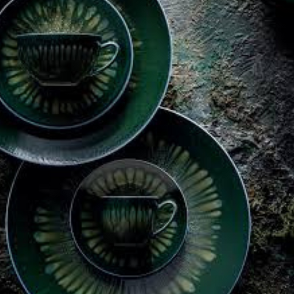 Two dark green teacups sit on matching Cyclone Dinnerware saucers and porcelain plates by Jaune De Chrome, arranged with overlapping edges. Decorative radial patterns and dramatic lighting highlight the textured surface.