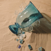 The Yakush Sal Blue Vase, a large hand-blown glass piece featuring decorative swirls, lies on its side in the sand with small fragments of crushed glass around its opening.