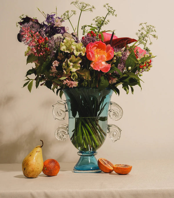 The Yakush Sal Blue Vase, hand-blown glass, is filled with colorful flowers on a beige tablecloth. In front are a pear, two tangerines, and a halved blood orange. The background features a plain light-colored wall.