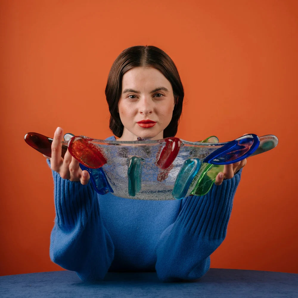 A woman in a blue sweater holds the Yakush Japanese Winter Bowl - Neon, a decorative bowl crafted from recycled blown glass with colorful glass accents, posing in front of an orange background and a blue table.