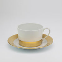 The Danielle Gold Dinnerware teacup by Royal Limoges features white porcelain with a gold band near the base and comes with a matching saucer adorned with a gold rim, shown on a plain light gray background.