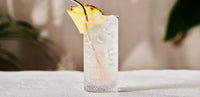 The Richard Brendon Diamond Highball, filled with ice and a clear drink, is garnished with a pineapple slice and copper straw on a white table, surrounded by cocktail glasses and a blurred green plant in the background.