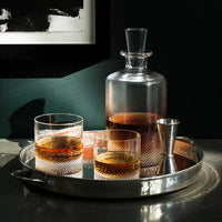 A glass decanter and two Richard Brendon Diamond DOF glasses rest on a round metal tray with a jigger, all reflecting light on a dark surface beneath framed art.