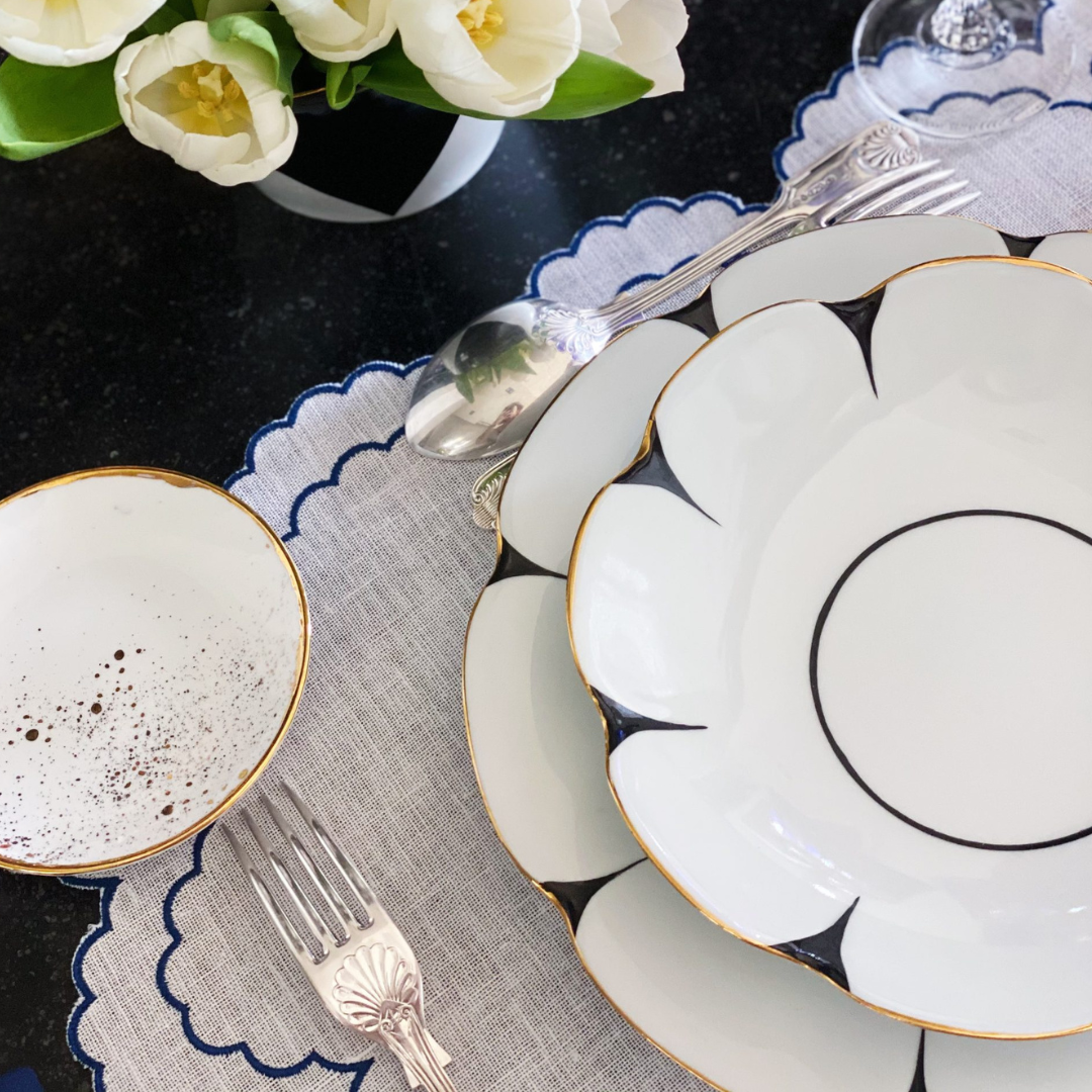 Elegant table setting with Molecot's Drops Dinnerware, gold-finished silverware, a white napkin on a scalloped gray placemat, a speckled bowl, and white flowers in a vase atop a dark surface.