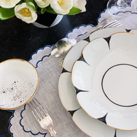 Elegant table setting with Molecot's Drops Dinnerware, gold-finished silverware, a white napkin on a scalloped gray placemat, a speckled bowl, and white flowers in a vase atop a dark surface.