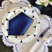 A table setting featuring Molecot's Drops Dinnerware—layered scalloped white plates—silver utensils, a blue napkin on top, and a gray placemat with blue trim. A white flower arrangement is partly visible in the corner.