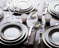 A formal dining table set with Bernardaud Dune Dinnerware, silverware, wine glasses, white cups, gray napkins, and small glass bowls of garnished appetizers on a reflective surface.