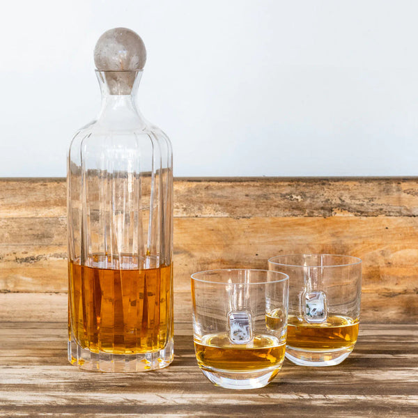 The Anna New York Elevo Smoke Agate Decanter, filled with amber liquid, sits on wood beside two matching whiskey glasses—each with a drink and a large die-shaped ice cube.