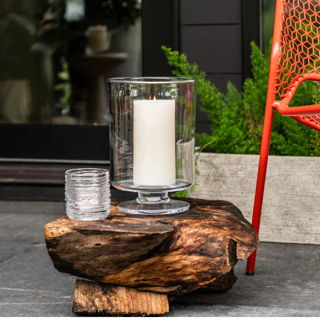 A lit Simon Pearce Echo Lake Tealight in Gift Box and an empty glass rest on a rustic wooden stump outdoors, surrounded by green plants and a red metal chair.