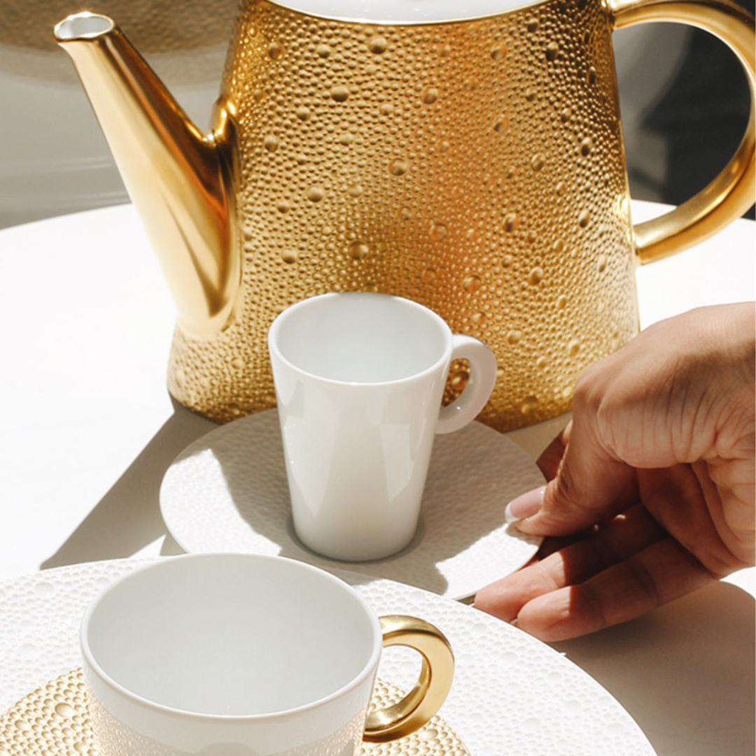 A hand reaches for a white cup on a textured saucer from Bernardaud’s Ecume Dinnerware Gold, beside another cup and a gold-accented teapot, all illuminated on a sunlit table.