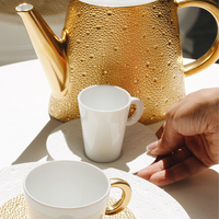 A hand reaches for a white cup on a textured saucer from Bernardaud’s Ecume Dinnerware Gold, beside another cup and a gold-accented teapot, all illuminated on a sunlit table.