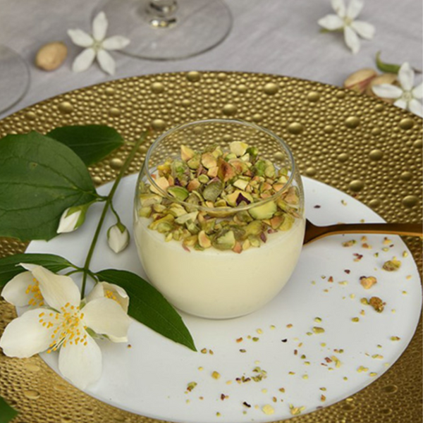 A glass of creamy dessert with chopped pistachios rests on Bernardaud's Ecume Dinnerware Gold, garnished with white flowers and green leaves, beside a gold spoon—an elegant display of contemporary style.