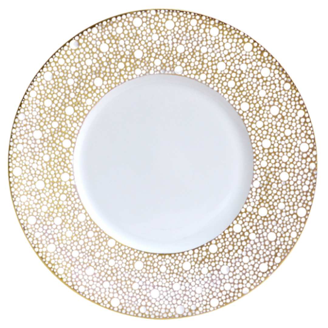 The Bernardaud Ecume Dinnerware Mordore is a round white porcelain plate with a wide rim accented by an intricate gold dotted pattern, while its center remains beautifully plain for a luxurious, elegant look.
