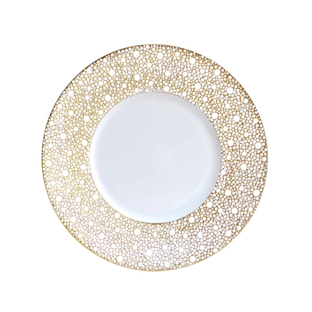 The Bernardaud Ecume Dinnerware Mordore is a white porcelain plate with a wide rim embellished by an intricate gold dotted circular pattern, while its center remains plain, highlighting the elegant design against the white background.