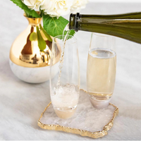 A bottle pours champagne into an Elevo Champagne Smoke Agate Glass by Anna New York, beside another filled glass. Both are on decorative coasters, next to a marble vase with white flowers and green leaves on a light surface.
